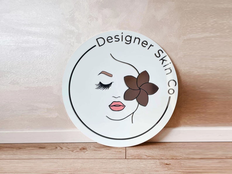 May include: White round sign with a black border and the text "Designer Skin Co."  A stylized illustration of a woman's face with closed eyes and a brown flower behind her eye.