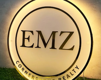 May include: A circular, illuminated sign featuring a white face with a black border. The letters EMZ are printed in large, bold black font in the center, with the text CORNERSTONE REALTY curved along the bottom edge in a smaller black font.