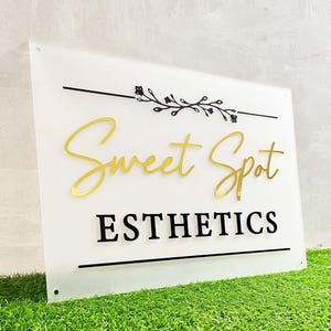 May include: White acrylic sign with black and gold lettering. The sign reads "Sweet Spot Esthetics" with a black floral design above the text.