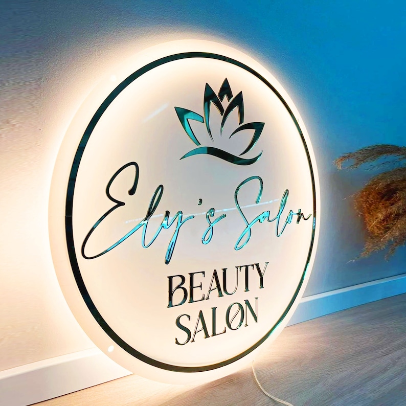 May include: A white round sign with a gold border and a lit halo. The sign has a stylized lotus flower design and the text "Ely's Salon Beauty Salon".