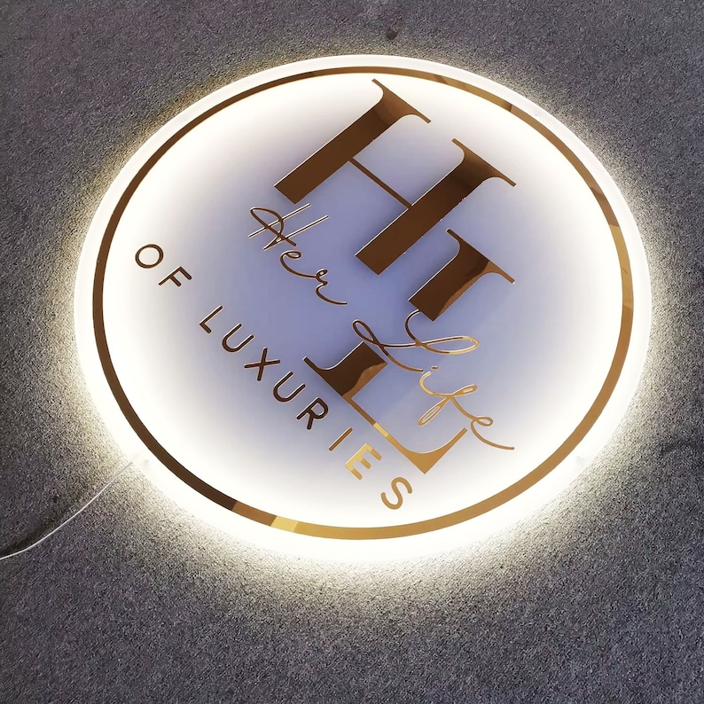 May include: A white circular sign with a gold outline and the text "Her Life of Luxuries" and the letters "HL" in gold.