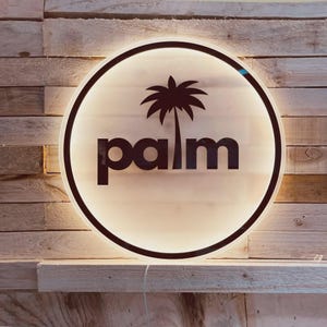May include: A round, illuminated sign with the word "palm" in a bold, black font, and a silhouette of a palm tree. The sign has a black border and is mounted on a wooden wall.