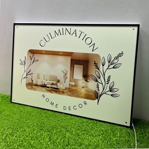 May include: A white rectangular sign with a black frame and black text that reads "CULMINATION HOME DECOR". The sign has a cutout in the centre that shows a picture of a living room with a sofa, coffee table, and plants. The sign is on a green surface.