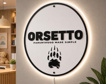 May include: A circular wall sign with a white face and black border, featuring the black text ORSETTO, the smaller text PARENTHOOD MADE SIMPLE, and a black bear paw print graphic. The sign is backlit with a soft white glow and has a power cord.