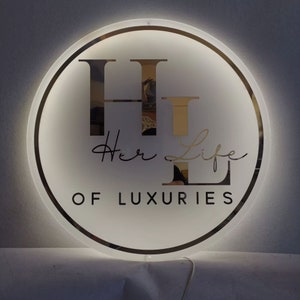 May include: A white circular sign with a black border and a gold-colored "H" and "L" in the center. The text "Her Life of Luxuries" is written in black below the letters.