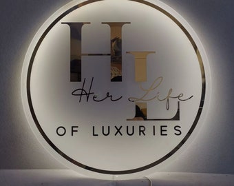 May include: A circular, backlit acrylic sign featuring a reflective gold finish. The design displays large letters H and L, the script text Her Life, and the block text OF LUXURIES. The sign emits a soft white glow from behind its perimeter.