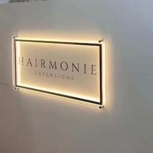 Op de afbeelding: Een wit rechthoekig bord met een zwarte lijst en een verlichte rand. Het bord heeft de tekst "Hairmonie Extensions" in zwarte letters.