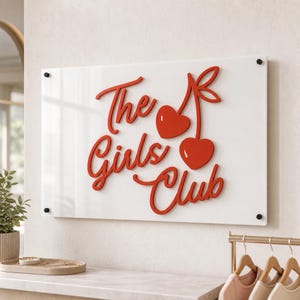 May include: A rectangular white acrylic sign featuring the red text The Girls Club in a cursive font, accompanied by a graphic of two red cherries. The sign is mounted with four black circular standoffs at each corner on a light-colored wall.