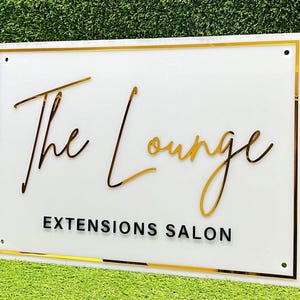 May include: White sign with a gold border and gold lettering that reads "The Lounge Extensions Salon".