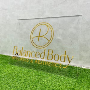 May include: A clear acrylic sign with gold lettering that reads "Balanced Body Wellness & Aesthetics". The sign features a gold circle with a stylized letter "B" inside.