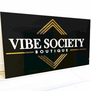 May include: A black sign with the words "VIBE SOCIETY BOUTIQUE" in white lettering. Gold geometric shapes and lines accent the text. The sign is rectangular and appears to be made of a glossy material.