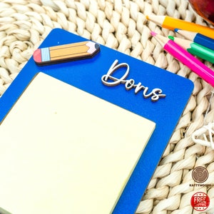 May include: Blue wooden notepad with a yellow sticky note, a wooden pencil, and the name "Dons" written in cursive on the front.