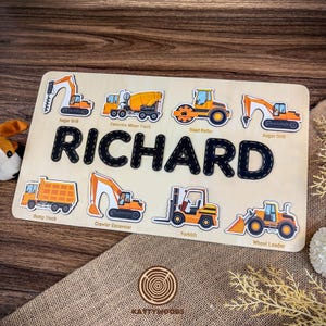 May include: A rectangular wooden puzzle board featuring the name RICHARD in large black letters. Surrounding the name are eight colorful illustrations of construction vehicles including an auger drill, concrete mixer, road roller, and forklift.