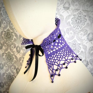 May include: A purple crocheted collar with black ribbon ties and black beads. The collar is on a white mannequin.