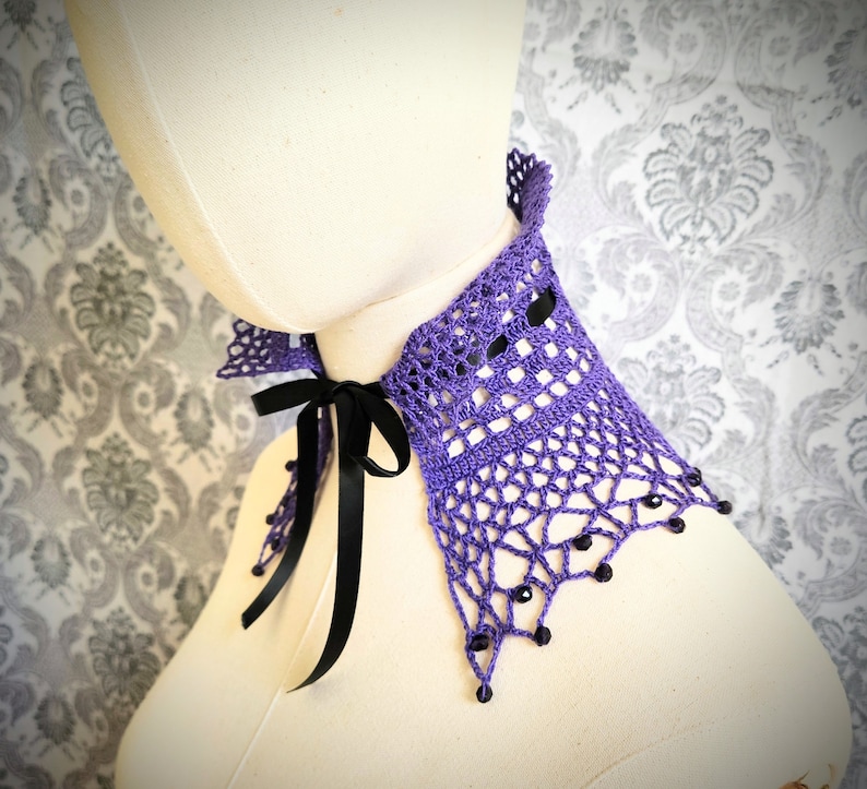 May include: A purple crocheted collar with black ribbon ties and black beads. The collar is on a white mannequin.