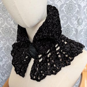 May include: A black velvet collar with a decorative button fastening. The collar has a scalloped edge and a textured, plush appearance. The button features a cameo design. The collar is displayed on a mannequin.