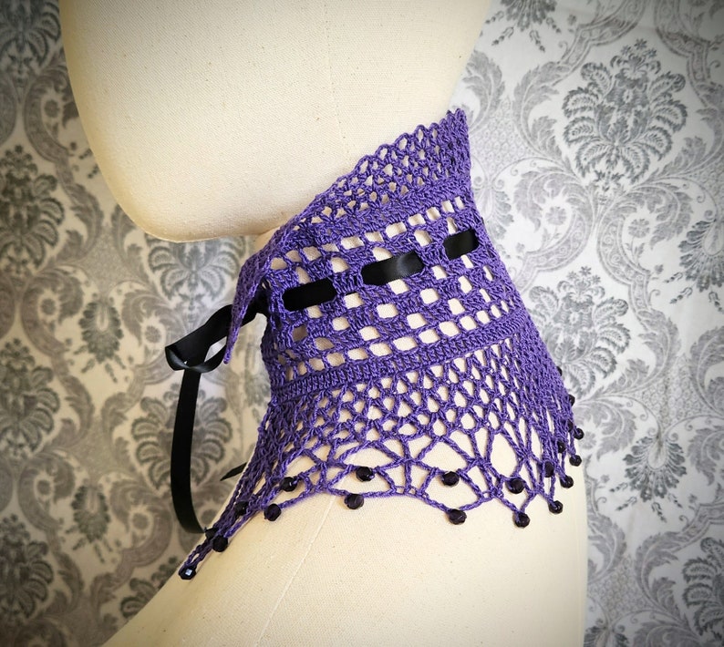 May include: Purple crochet collar with black ribbon and black beads. The collar is on a white mannequin.