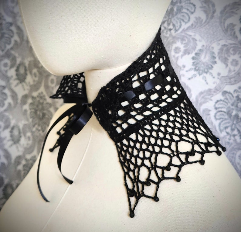 May include: Black crocheted lace choker with a black satin ribbon tie. The choker has a delicate, intricate design and is adorned with small black beads.