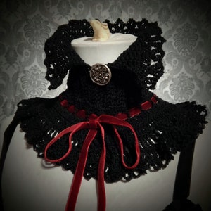 May include: Black crocheted collar with a red velvet ribbon tied in a bow and a silver button in the center.
