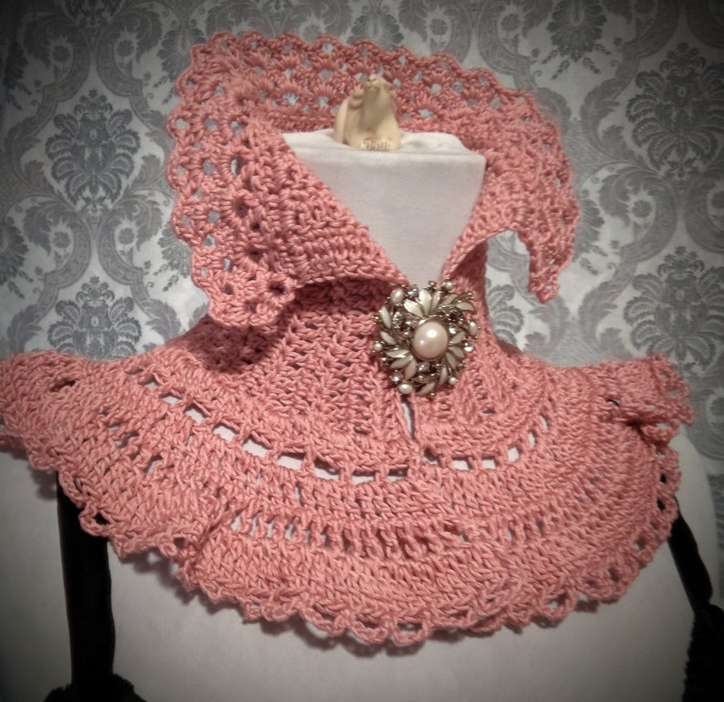 May include: A pink crocheted collar with a decorative brooch. The collar has a scalloped edge and is made with a delicate stitch.