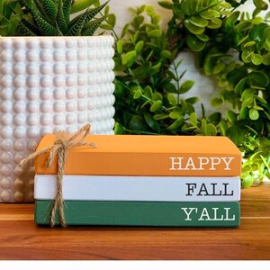 May include: Stacked decorative books in orange, white, and green, tied with twine. The books read "HAPPY FALL Y'ALL" in white lettering. A white dotted planter with a succulent and a green wreath are in the background.