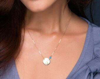 Puede incluir: Un collar de oro con un colgante redondo y una pequeña cuenta azul turquesa. El collar lo lleva una persona con el pelo largo y castaño.