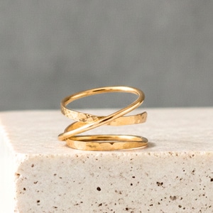 May include: A gold-colored ring featuring a multi-layered, crossover design. The band consists of three distinct, thin, overlapping loops that create a spiral effect. The metal surface has a subtle, hammered texture throughout the entire piece.