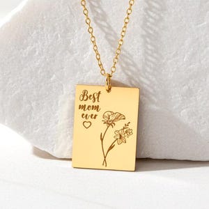 May include: A gold-colored pendant necklace with a rectangular charm. The charm has the words "Best mom ever" and a floral design engraved on it. The necklace is displayed on a white, textured surface.