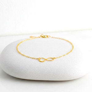 May include: A delicate gold bracelet with an infinity symbol at its centre. The bracelet is made of a fine chain and is displayed on a white, rounded surface. The infinity symbol is a continuous loop, symbolising eternity.