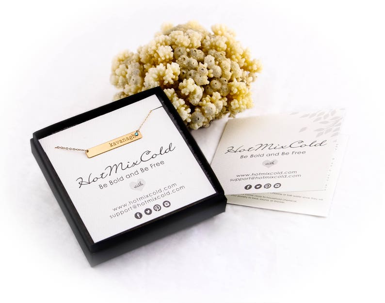 Puede incluir: Un collar de barra de oro con el nombre "kavanagh" grabado. El collar est&aacute; en una caja negra con las palabras "Hot Mix Cold" y "Be Bold and Be Free" en ella. La caja est&aacute; sobre una superficie blanca.