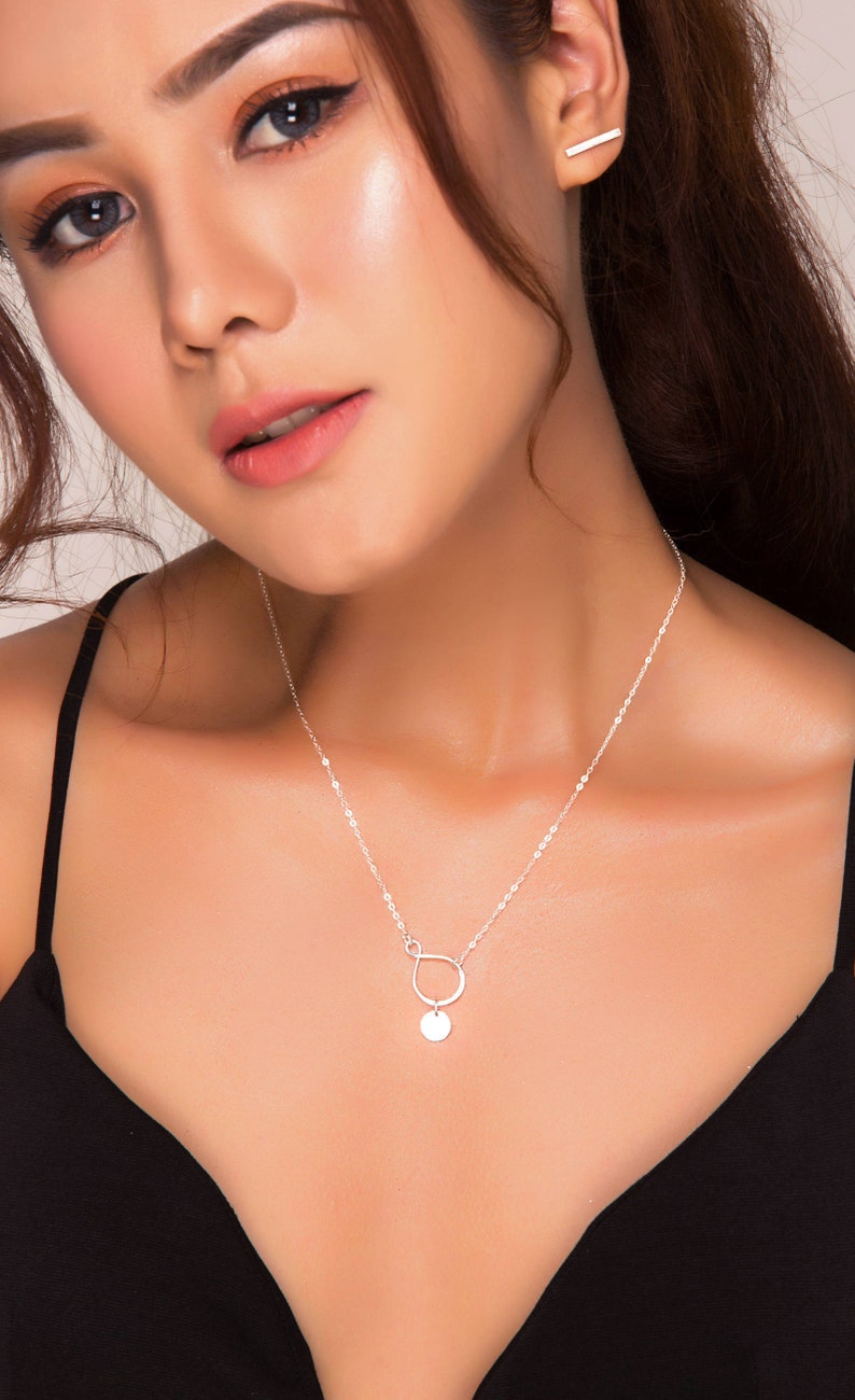 Puede incluir: Un collar de plata con una cadena delicada y un colgante con un c&iacute;rculo peque&ntilde;o y una forma curva. El collar lo lleva una persona con el pelo oscuro y una blusa negra.
