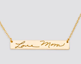 May include: Gold-colored bar necklace with the inscription "Love Mom" in a cursive font. The pendant is rectangular and suspended from a delicate chain. The necklace is displayed against a light gray background.