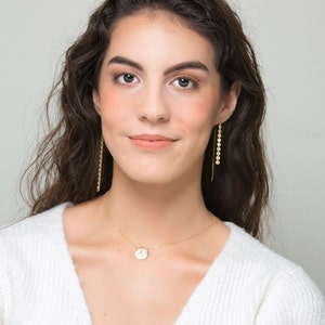 K&ouml;nnte beinhalten: Eine Frau mit langen braunen Haaren tr&auml;gt einen wei&szlig;en Pullover und eine goldene Halskette mit einem runden Anh&auml;nger. Sie tr&auml;gt goldene Ohrringe mit kleinen runden Anh&auml;ngern.