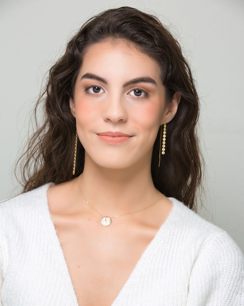 K&ouml;nnte beinhalten: Eine Frau mit langen braunen Haaren tr&auml;gt einen wei&szlig;en Pullover und eine goldene Halskette mit einem runden Anh&auml;nger. Sie tr&auml;gt goldene Ohrringe mit kleinen runden Anh&auml;ngern.