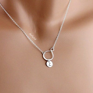 Puede incluir: Un collar de plata con una cadena delicada y un colgante en forma de s&iacute;mbolo de infinito invertido. El colgante tiene un peque&ntilde;o c&iacute;rculo unido a &eacute;l con la letra "L" grabada en &eacute;l.