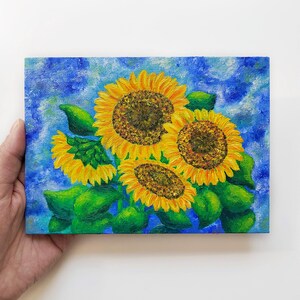 以下が含まれることがあります： ひまわりの長方形の絵。このアートワークは、黄色の花びらと茶色の中心を持つ4つのひまわりを特徴とし、緑の葉に囲まれています。背景は、白いアクセントのあるテクスチャードブルーです。