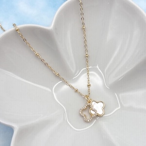 Op de afbeelding: Een delicate goudkleurige ketting met een klavervormige hanger. Eén klaver is omlijnd met kleine, heldere kristallen, en de andere is van wit, parelmoerachtig materiaal. De ketting ligt op een witte, bloemvormige schaal tegen een lichtblauwe achtergrond.