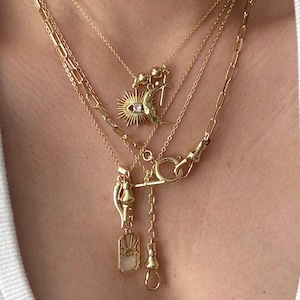 May include: A collection of layered gold necklaces. The necklaces feature various pendants, including a sun with an eye, a crescent moon, a cross, a horn, and a rectangular pendant. The chains have different styles, including a paperclip chain and delicate link chains.