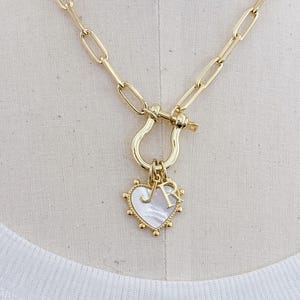May include: Gold-toned necklace with a heart-shaped pendant. The pendant features a mother-of-pearl inlay and the letters "J" and "R". The necklace has a large link chain and a decorative clasp. The heart pendant is approximately 1 inch in width.