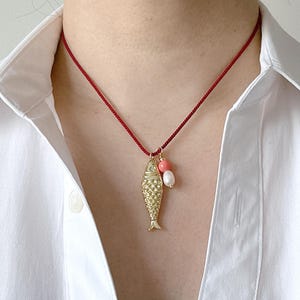 Op de afbeelding: Goudkleurige vishangerketting aan een rode koord. De vis heeft een textuur met schubben en is gecombineerd met een koraal kraal en een witte parel. De ketting wordt gedragen over een wit overhemd.