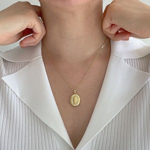May include: A gold-toned oval pendant necklace featuring a central embossed figure surrounded by a border of small clear stones. The pendant hangs from a delicate gold-toned chain, resting against a white ribbed collared shirt.