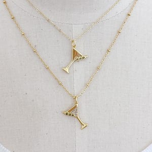 Peut inclure: Deux colliers dorés avec des pendentifs en forme de verre à martini. Les pendentifs ont un design triangulaire avec de petits accents verts et clairs. Les colliers sont sur fond blanc.