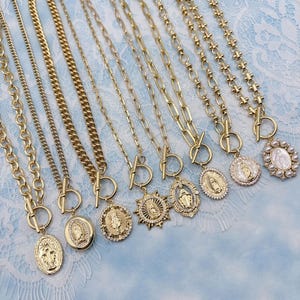 May include: A selection of gold-coloured necklaces with diverse chain designs and pendants. Some pendants display religious iconography, such as the Virgin Mary, and are embellished with crystals or pearls. The necklaces are presented on a light blue lace background.