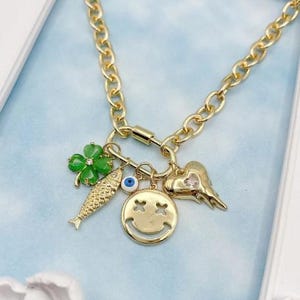 May include: Gold chain necklace with a variety of charms, including a green four-leaf clover, a blue evil eye, a gold fish, a heart with a rhinestone, and a gold smiley face.
