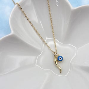 May include: A gold necklace with a fine chain, featuring a pendant with a blue evil eye charm and a gold horn. The necklace is presented on a white, petal-shaped dish.