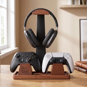 May include: A black and walnut wood gaming organizer stand featuring a central vertical post for headphones, two controller cradles, and a rectangular base. The left controller cradle is engraved with the text Best Dad in a serif font.