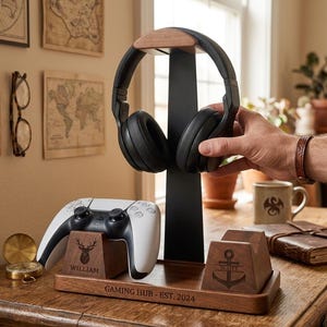 May include: A wooden gaming organizer stand featuring a central black vertical headphone holder, two controller docks with engraved deer and anchor icons, and the text "GAMING HUB - EST. 2024" on the base, holding black headphones and a white controller.