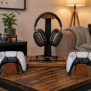 May include: A black metal headphone stand with a wooden base, holding dark gray over-ear headphones. Two white and black wireless game controllers rest on individual wooden stands, with a pair of black-rimmed glasses placed on the central base.