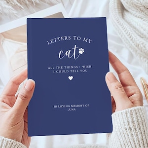 May include: A navy blue book with the title "Letters to My Cat" in white text, featuring a paw print. The book also includes the text "All the things I wish I could tell you" and "In loving memory of Luna". A gold pen rests on the white surface.