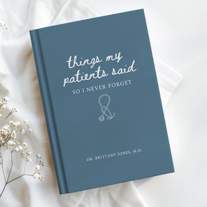 Puede incluir: Un diario azul con el texto "things my patients said so I never forget" y un gráfico de estetoscopio. El diario está sobre una superficie blanca con flores blancas.